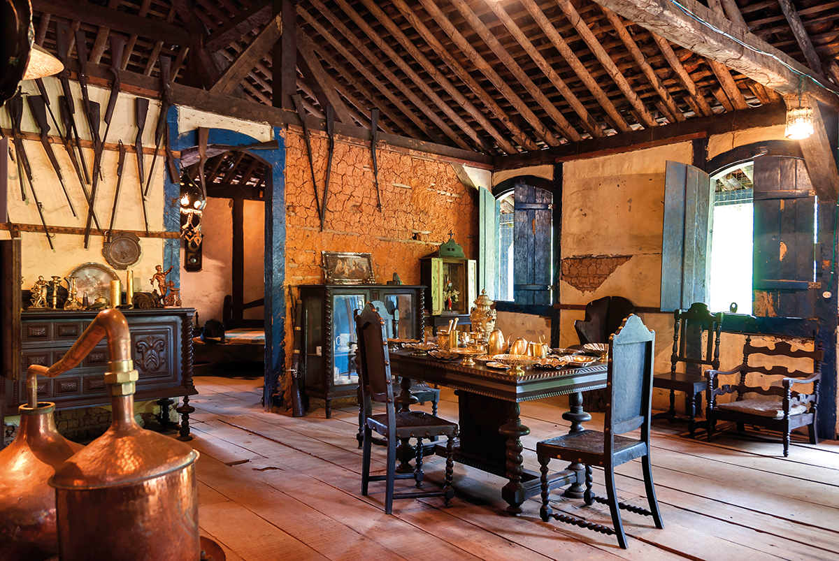 sala de uma casa antiga com teto de madeira paredes de barro com armas penduradas, no centro uma mesa retangular com 3 cadeiras e ao redor móveis antigos espalhados