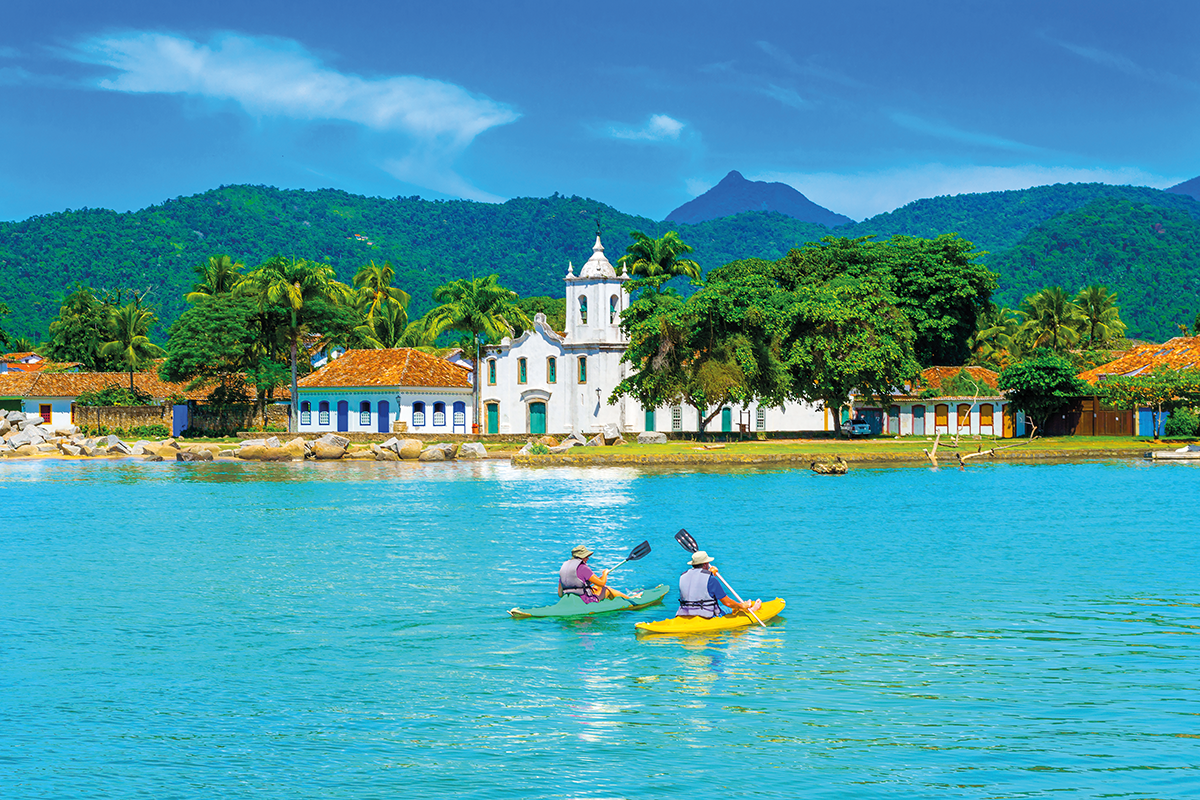 duas pessoas passeando em caiaques (um verde e um amarelo) na água azul e ao fundo uma igreja e arvores