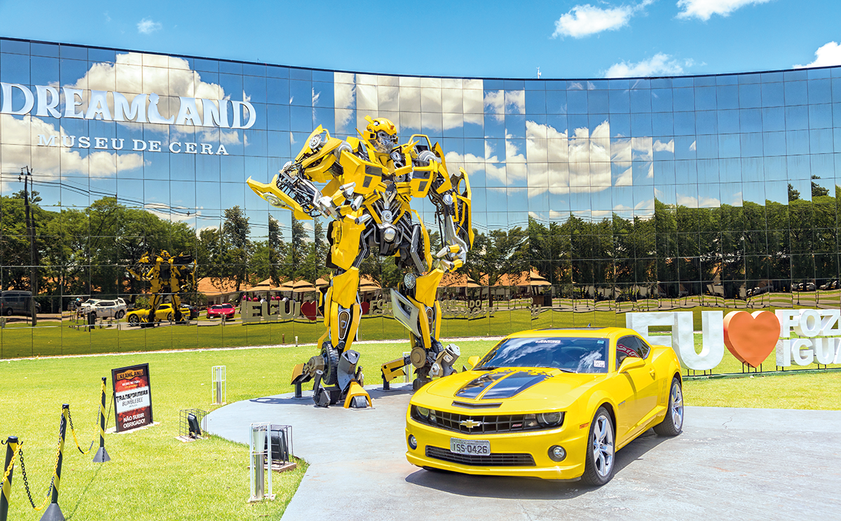 robô transformer amarelo bumblle bee e a ao lado sua versão carro, atrás um prédio espelhado