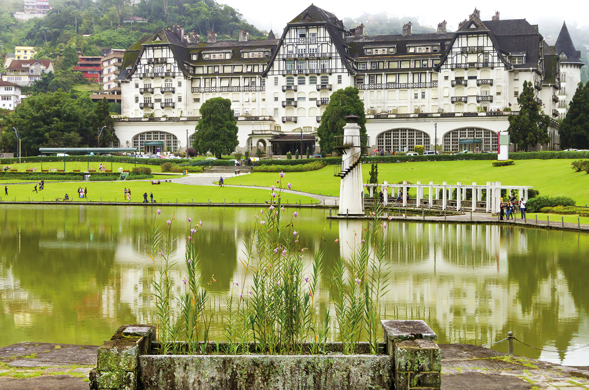 Em primeiro plano, um lago com água esverdeada, ao fundo, pessoas em escala bem pequena em frente a um gigantesco prédio branco, de arquitetura colonial